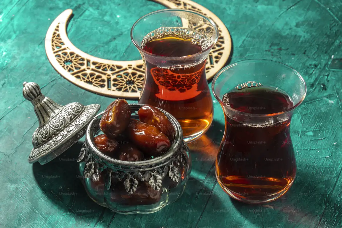 Zwei Gläser Schwarztee neben einer Schale mit Datteln auf türkisfarbenem Grund.