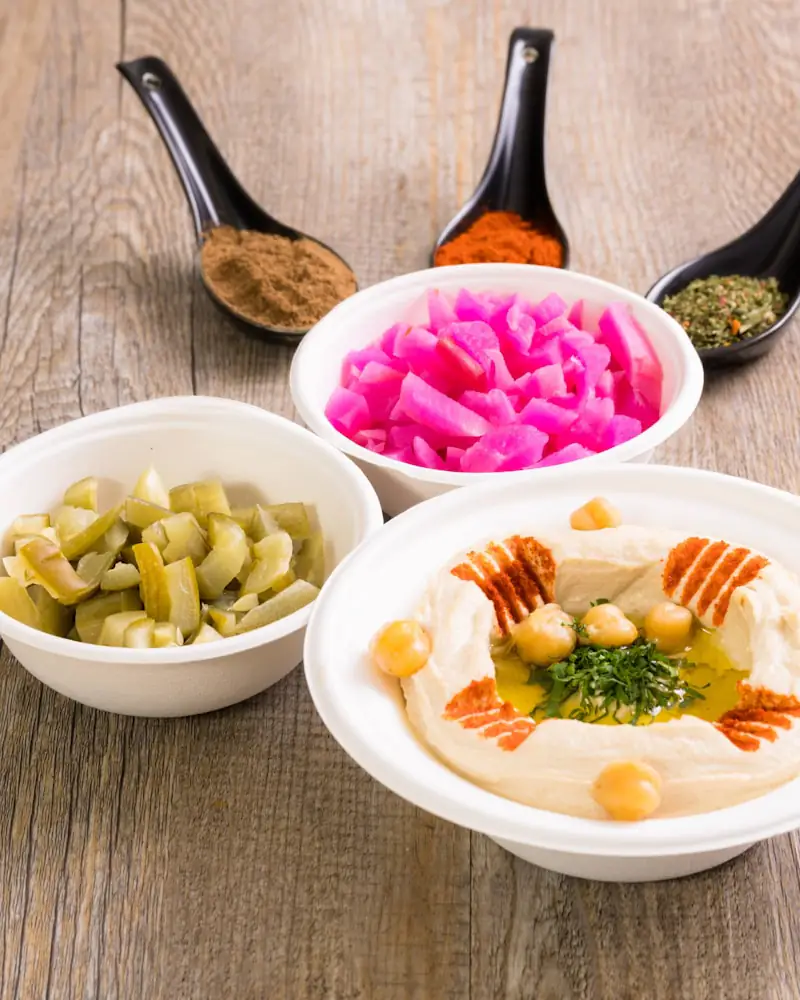 Drei weiße Schalen nebeneinander mit Hummus, eingelegtem Gemüse und Kräutern.